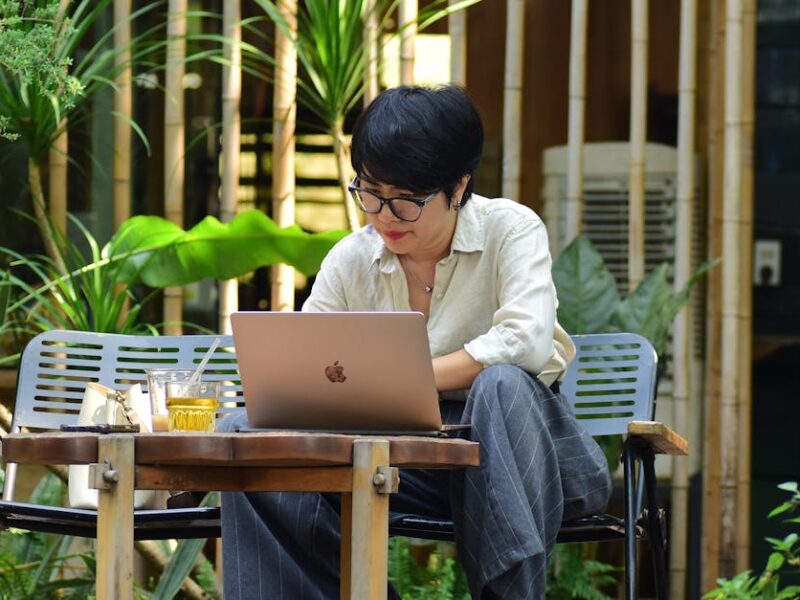A woman focuses on her laptop while sitting in a lush outdoor cafe.