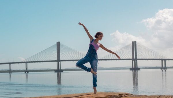 A woman performs a yoga pose on rocks near a scenic bridge over calm waters.