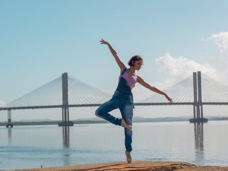 A woman performs a yoga pose on rocks near a scenic bridge over calm waters.