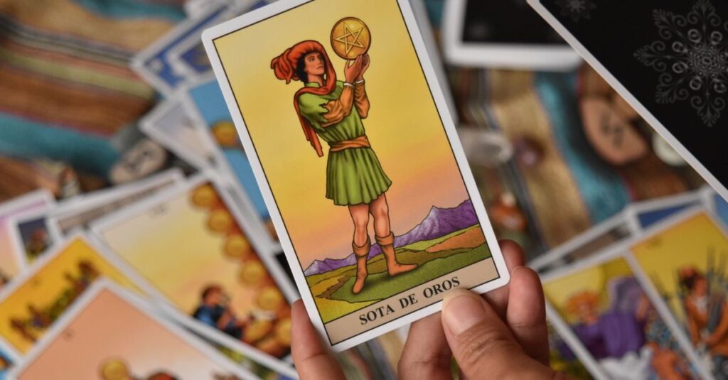 A person holds the Sota de Oros tarot card among a spread of cards for a reading.