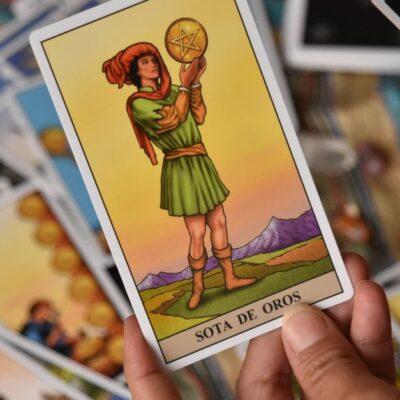 A person holds the Sota de Oros tarot card among a spread of cards for a reading.