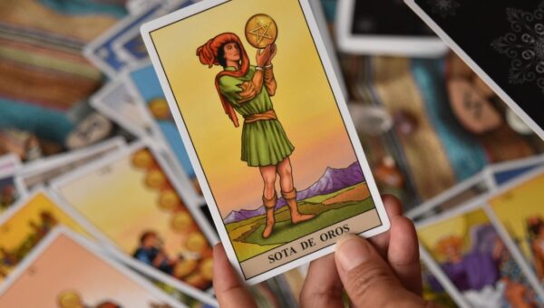 A person holds the Sota de Oros tarot card among a spread of cards for a reading.