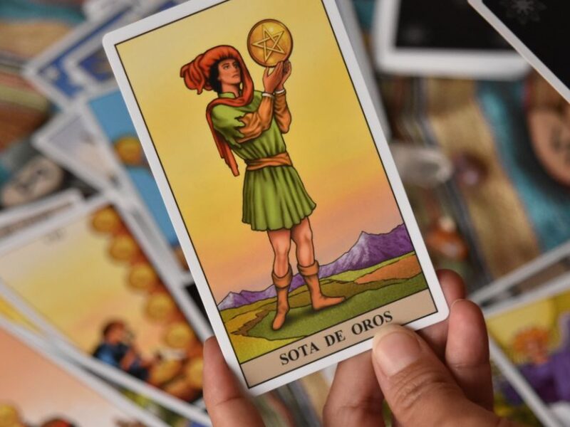 A person holds the Sota de Oros tarot card among a spread of cards for a reading.