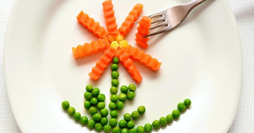 5 Maneiras de Usar a Criatividade na Alimentação Saudável 2 A playful flower made from carrots and peas on a white plate with a fork, showcasing creativity in food art.