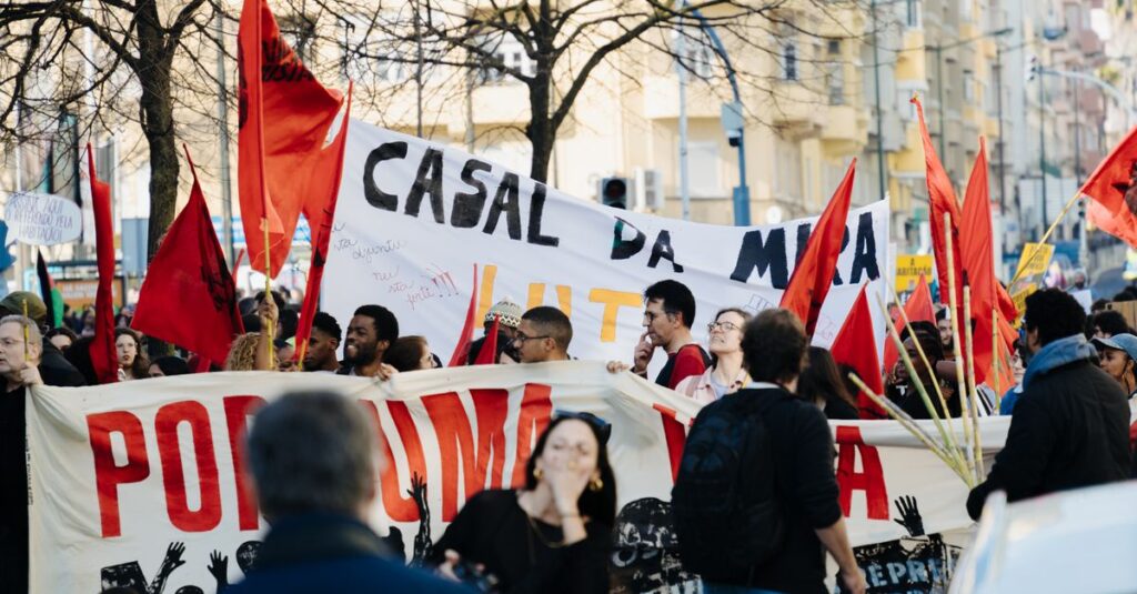 Group of people in a city staging a protest with banners and red flags during the day.