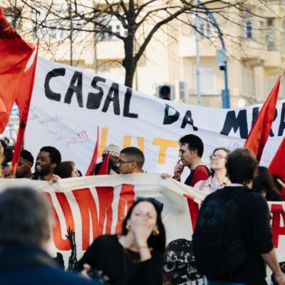 Group of people in a city staging a protest with banners and red flags during the day.