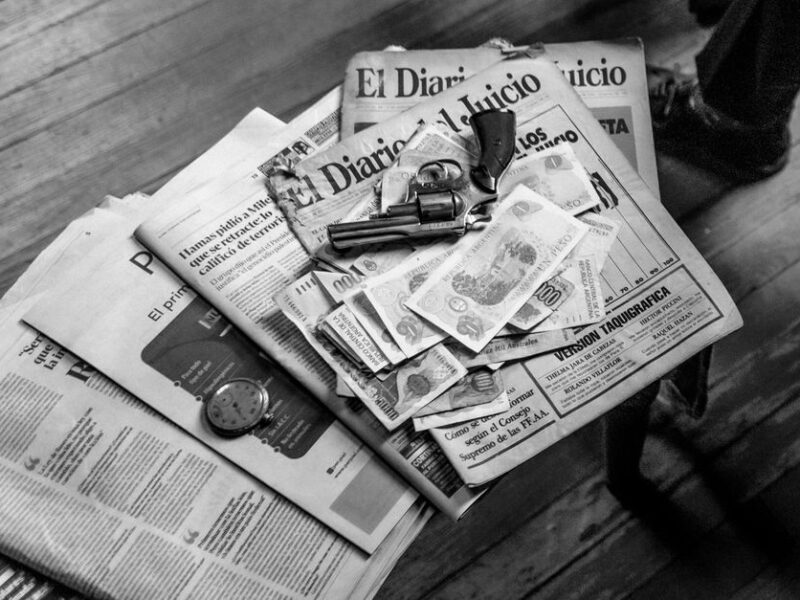 Black and white image featuring currency, newspapers, and a revolver on a wooden surface.