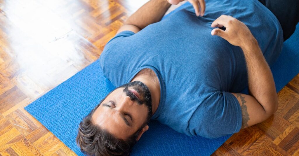 Adult man lying on a blue yoga mat indoors with eyes closed, focusing on relaxation and mindfulness.