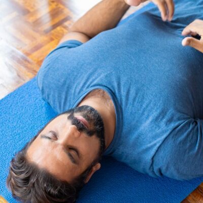 Adult man lying on a blue yoga mat indoors with eyes closed, focusing on relaxation and mindfulness.