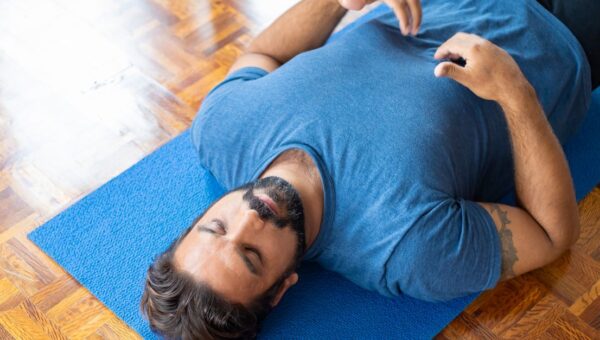Adult man lying on a blue yoga mat indoors with eyes closed, focusing on relaxation and mindfulness.