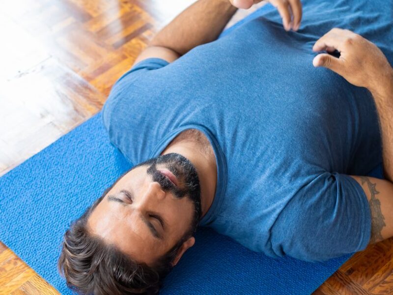 Adult man lying on a blue yoga mat indoors with eyes closed, focusing on relaxation and mindfulness.