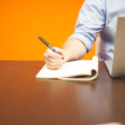 Adult writing notes in a notebook beside a laptop on a wooden desk with orange background.