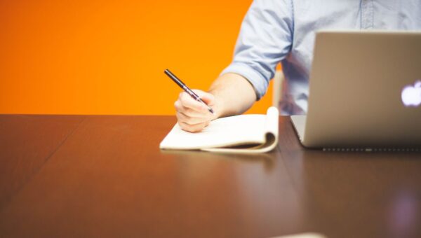 Adult writing notes in a notebook beside a laptop on a wooden desk with orange background.