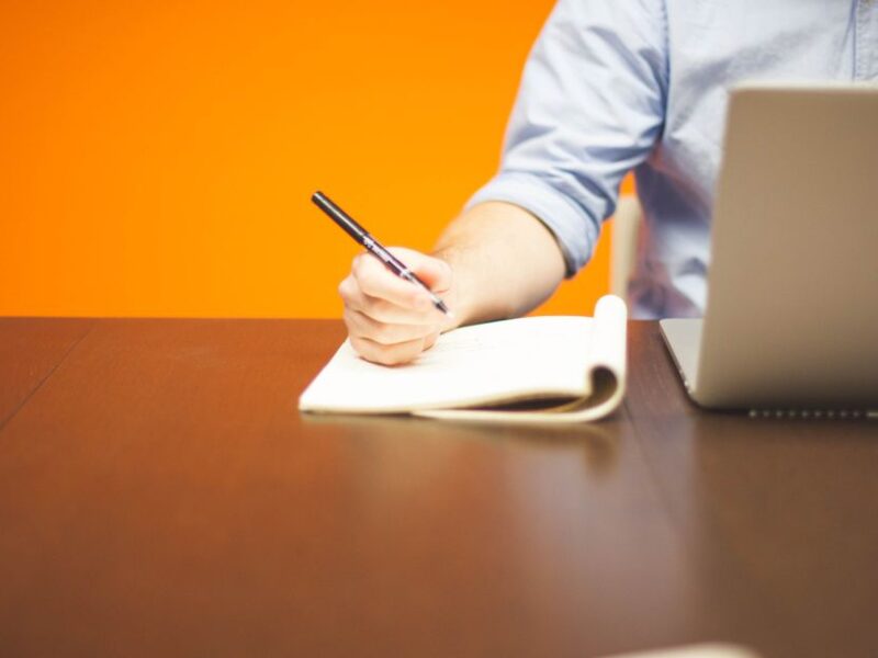 Adult writing notes in a notebook beside a laptop on a wooden desk with orange background.