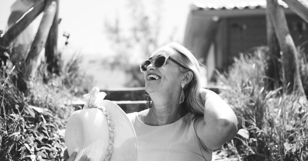 A smiling senior woman enjoys a sunny day outdoors wearing sunglasses and a sunhat.