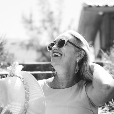 A smiling senior woman enjoys a sunny day outdoors wearing sunglasses and a sunhat.