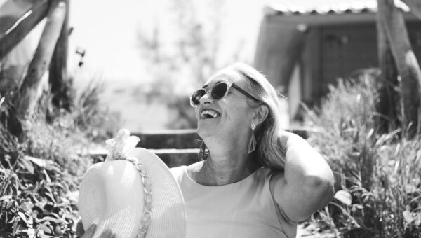 A smiling senior woman enjoys a sunny day outdoors wearing sunglasses and a sunhat.