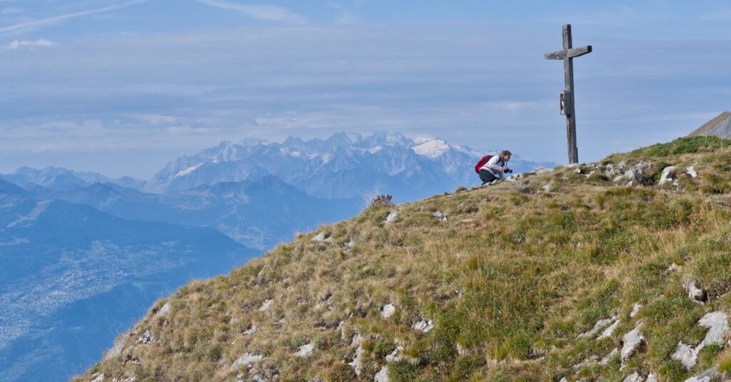 A hiker pauses at a mountain peak marked by a cross, overlooking stunning Alpine views.