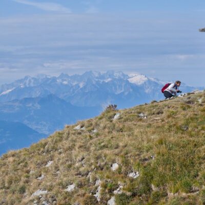 A hiker pauses at a mountain peak marked by a cross, overlooking stunning Alpine views.