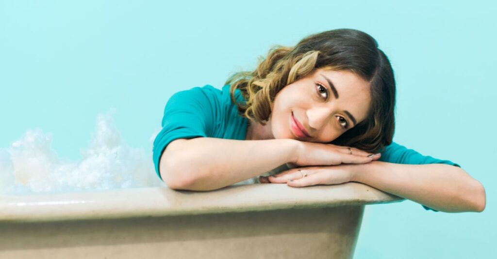 Woman with brown hair smiling and leaning on bathtub with blue background.