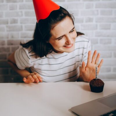 Woman in party hat waving at laptop and cupcake, celebrating virtual birthday.