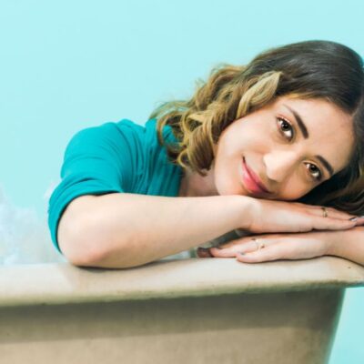 Woman with brown hair smiling and leaning on bathtub with blue background.