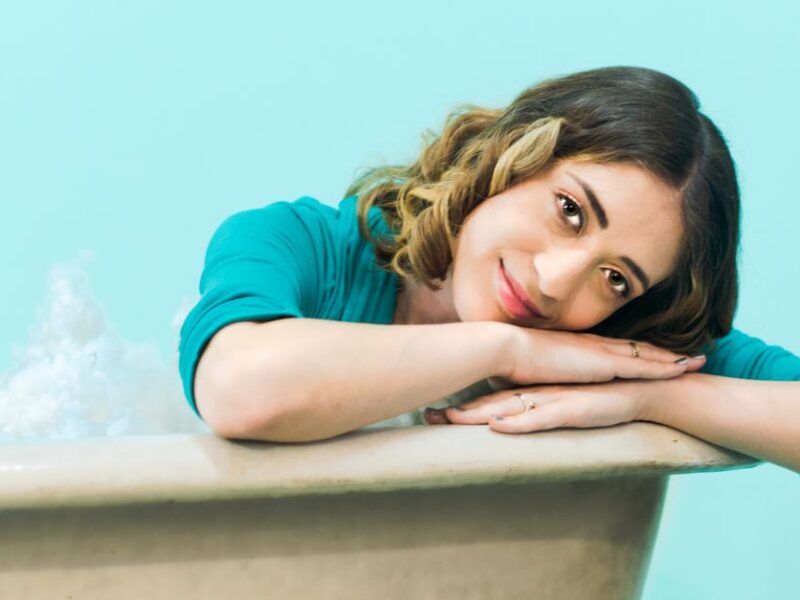 Woman with brown hair smiling and leaning on bathtub with blue background.