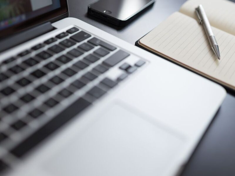 A modern workspace featuring a laptop, notebook, smartphone, and pen on a desk.