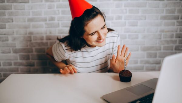 Woman in party hat waving at laptop and cupcake, celebrating virtual birthday.