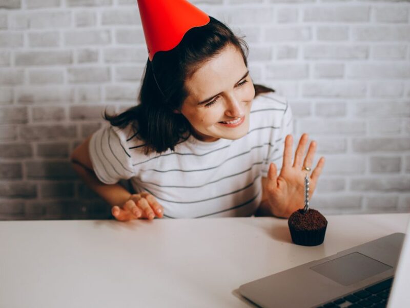Woman in party hat waving at laptop and cupcake, celebrating virtual birthday.