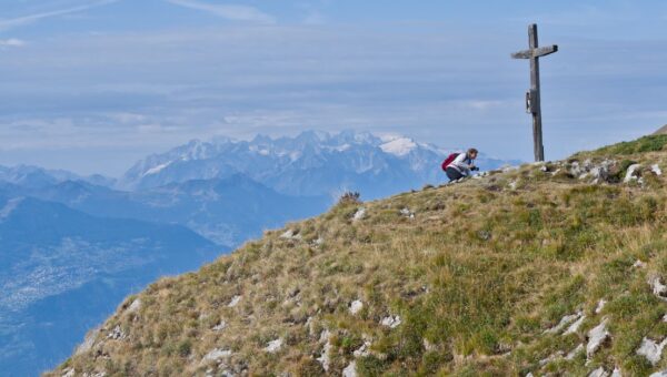 A hiker pauses at a mountain peak marked by a cross, overlooking stunning Alpine views.