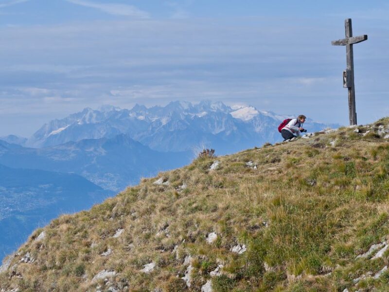 A hiker pauses at a mountain peak marked by a cross, overlooking stunning Alpine views.