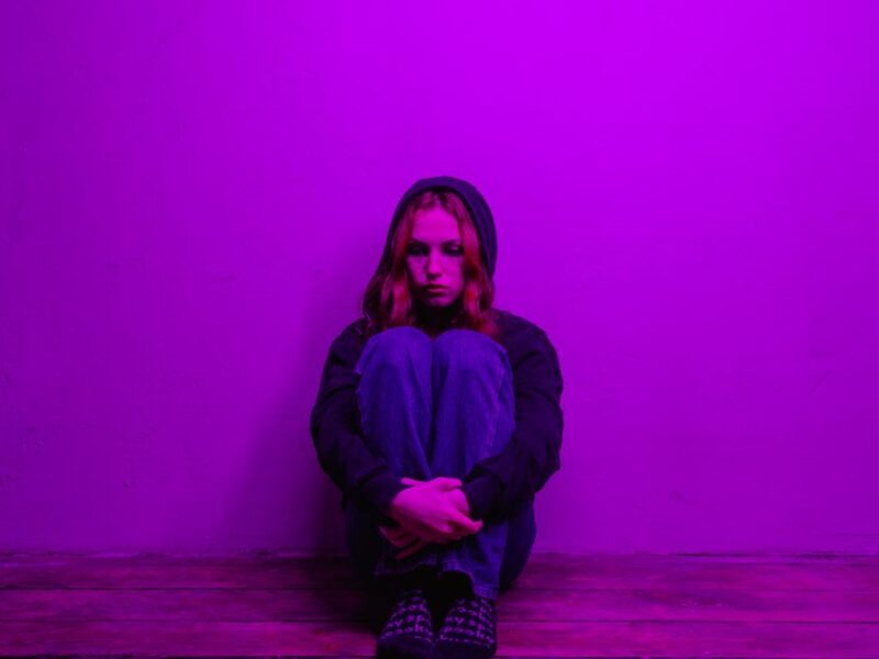 A woman in a hoodie sits alone on the floor, illuminated by purple light indoors.