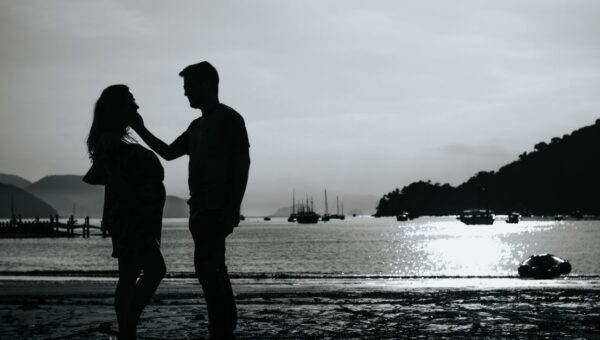 Black and white full body side view of anonymous couple caressing on shore near sea with various boats against pier