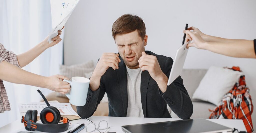 Adult man stressing out at home office desk with multiple demands surrounding him.