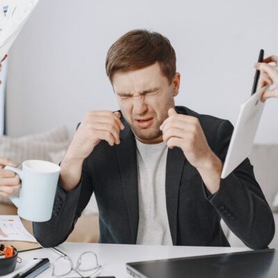 Adult man stressing out at home office desk with multiple demands surrounding him.