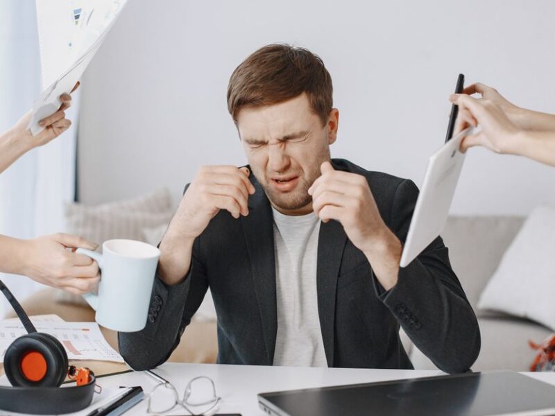 Adult man stressing out at home office desk with multiple demands surrounding him.