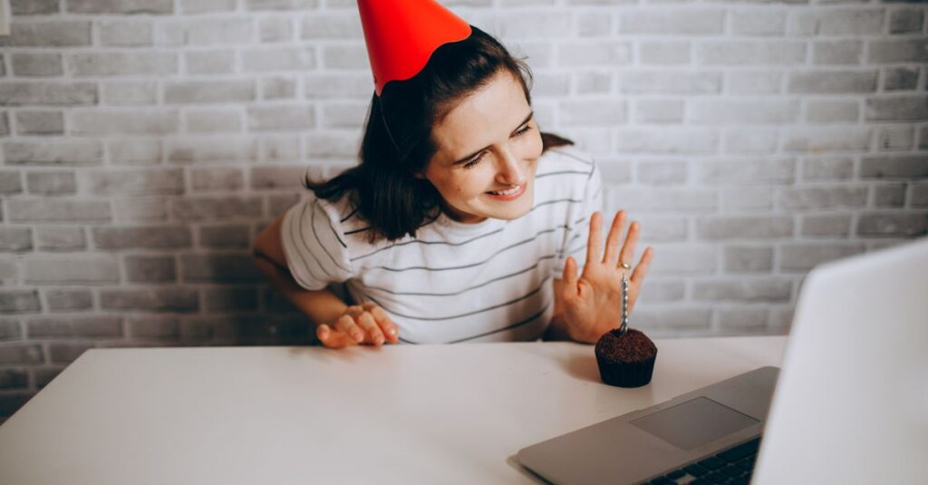 5 Dicas para Organizar um Encontro Virtual Incrível com Amigas 2 Woman in party hat waving at laptop and cupcake, celebrating virtual birthday.