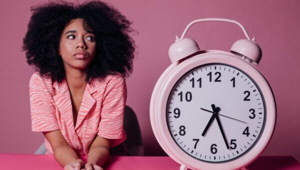 Pensive woman with afro hair leaning on a table next to a large pink alarm clock.