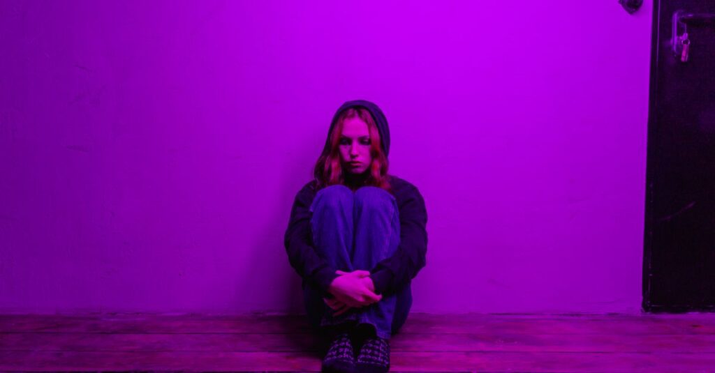 A woman in a hoodie sits alone on the floor, illuminated by purple light indoors.