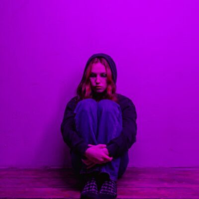A woman in a hoodie sits alone on the floor, illuminated by purple light indoors.