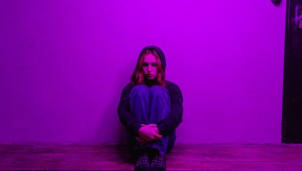 A woman in a hoodie sits alone on the floor, illuminated by purple light indoors.