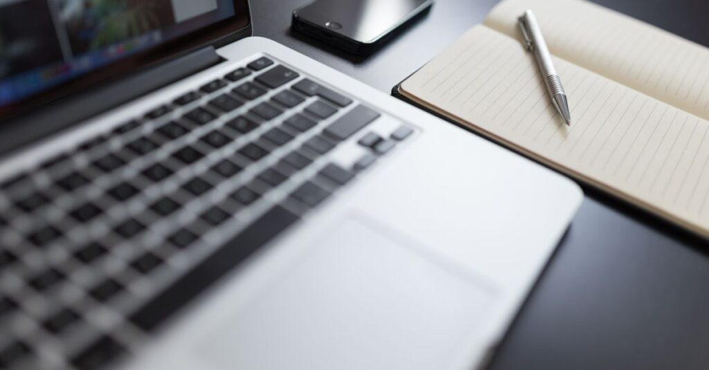 5 Dicas para Criar um Espaço de Trabalho Inspirador em Casa 2 A modern workspace featuring a laptop, notebook, smartphone, and pen on a desk.