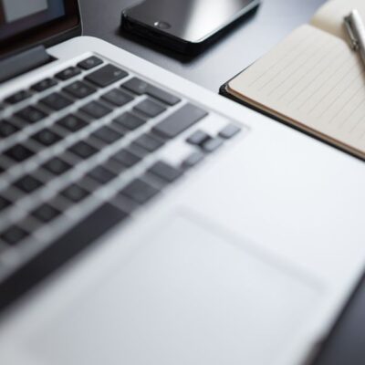 A modern workspace featuring a laptop, notebook, smartphone, and pen on a desk.