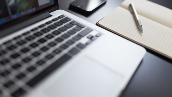 A modern workspace featuring a laptop, notebook, smartphone, and pen on a desk.