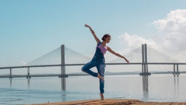 A woman performs a yoga pose on rocks near a scenic bridge over calm waters.
