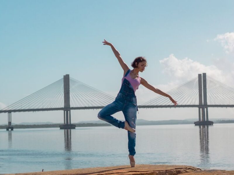 A woman performs a yoga pose on rocks near a scenic bridge over calm waters.