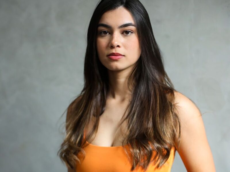 Elegant portrait of a young woman with long, dark hair in a vibrant orange top against a neutral background.