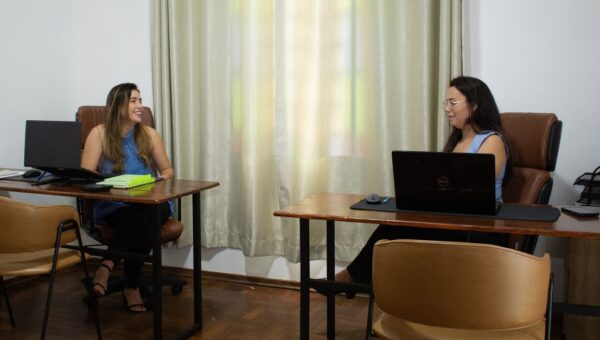 Two businesswomen in an office discussing work across desks with laptops.