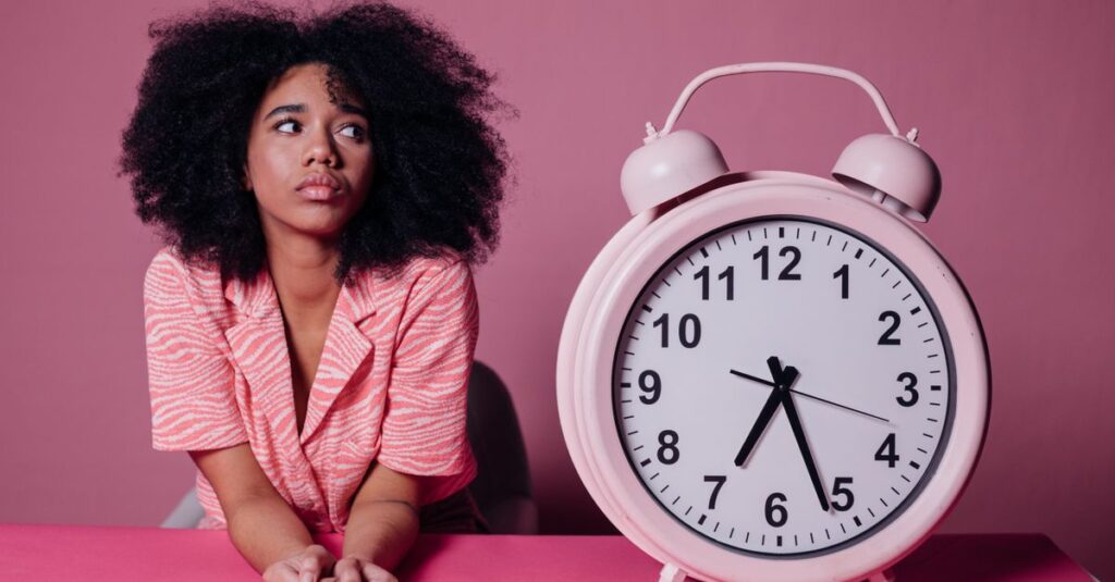 Como Lidar com a Procrastinação e Aumentar sua Criatividade 2 Pensive woman with afro hair leaning on a table next to a large pink alarm clock.